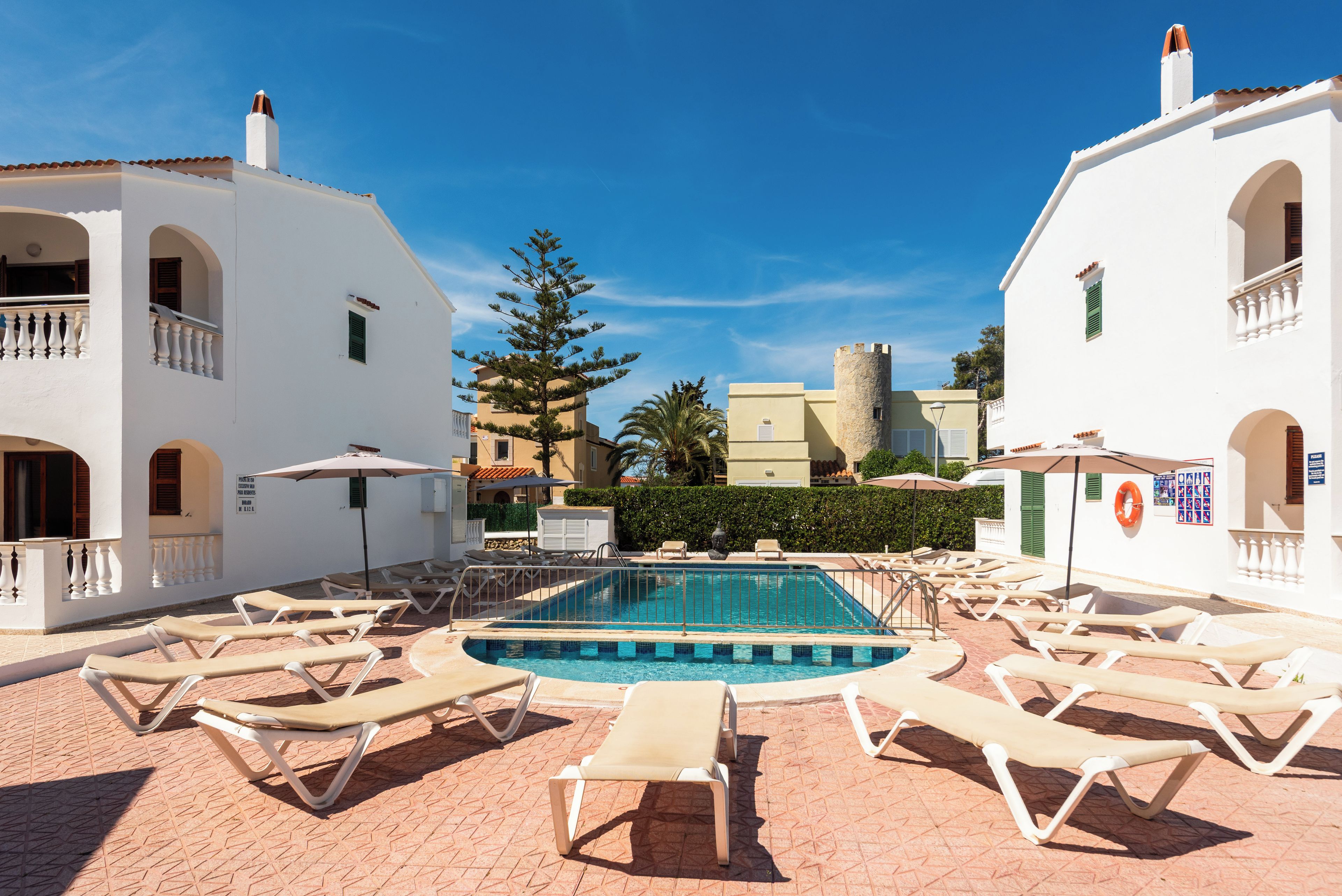 A pool area with sun loungers, a clear blue sky, and white buildings with balconies.