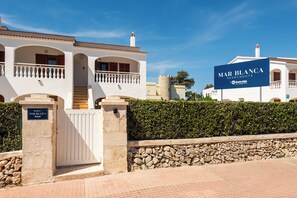 Una entrada con reja a Mar Blanca Apartments, con un muro de piedra y un cielo azul claro.