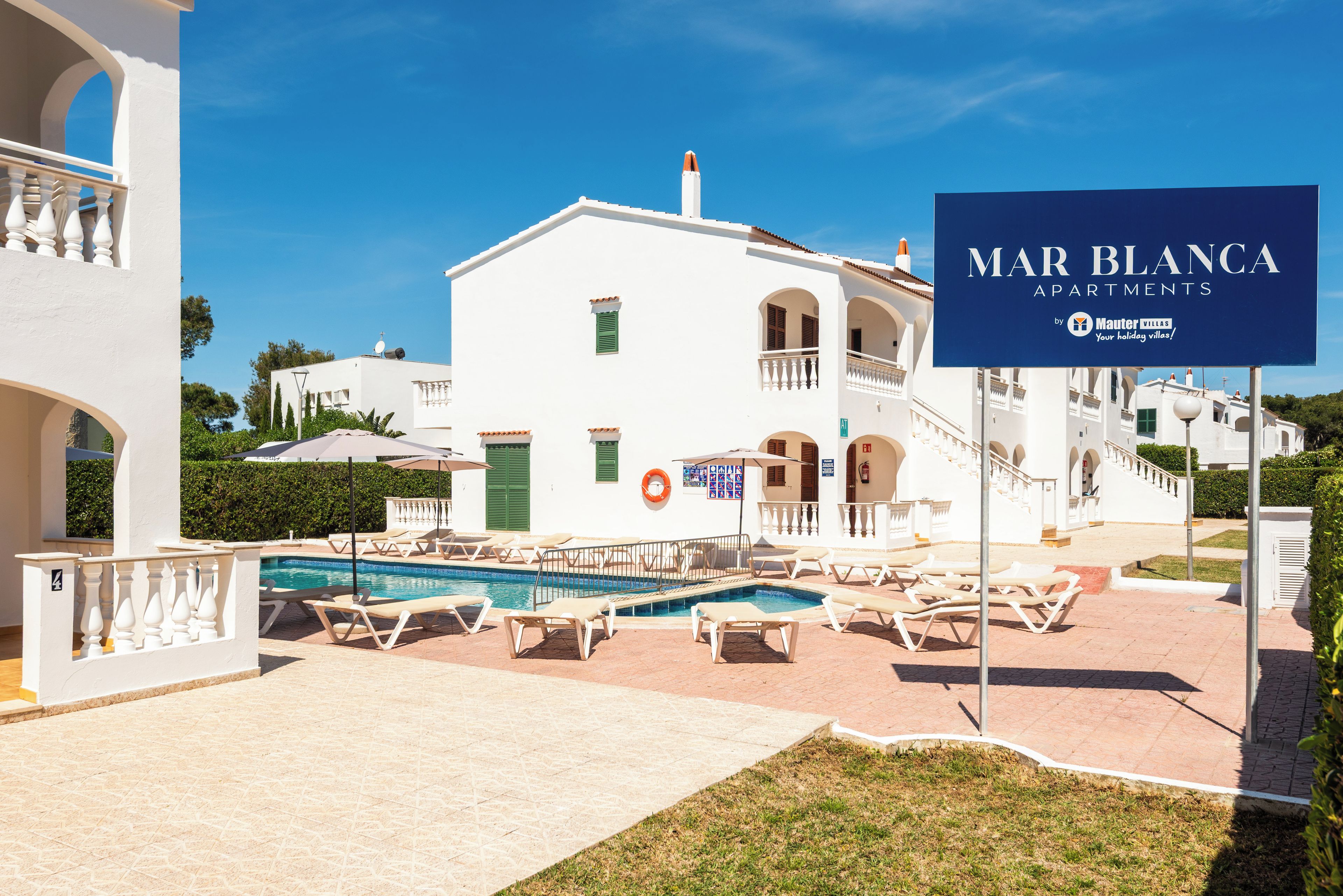En hvid bygning med swimmingpool, udendørs siddepladser og et skilt med teksten Mar Blanca Apartments.