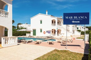 A white building with a swimming pool, outdoor seating, and a sign for Mar Blanca Apartments. - Apartamentos Mar Blanca (Ciutadella de Menorca)