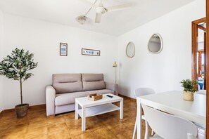 A living room with a sofa, a coffee table, a dining table, a ceiling fan, a wall-mounted mirror, and a potted plant. - Apartamentos Mar Blanca (Ciutadella de Menorca)