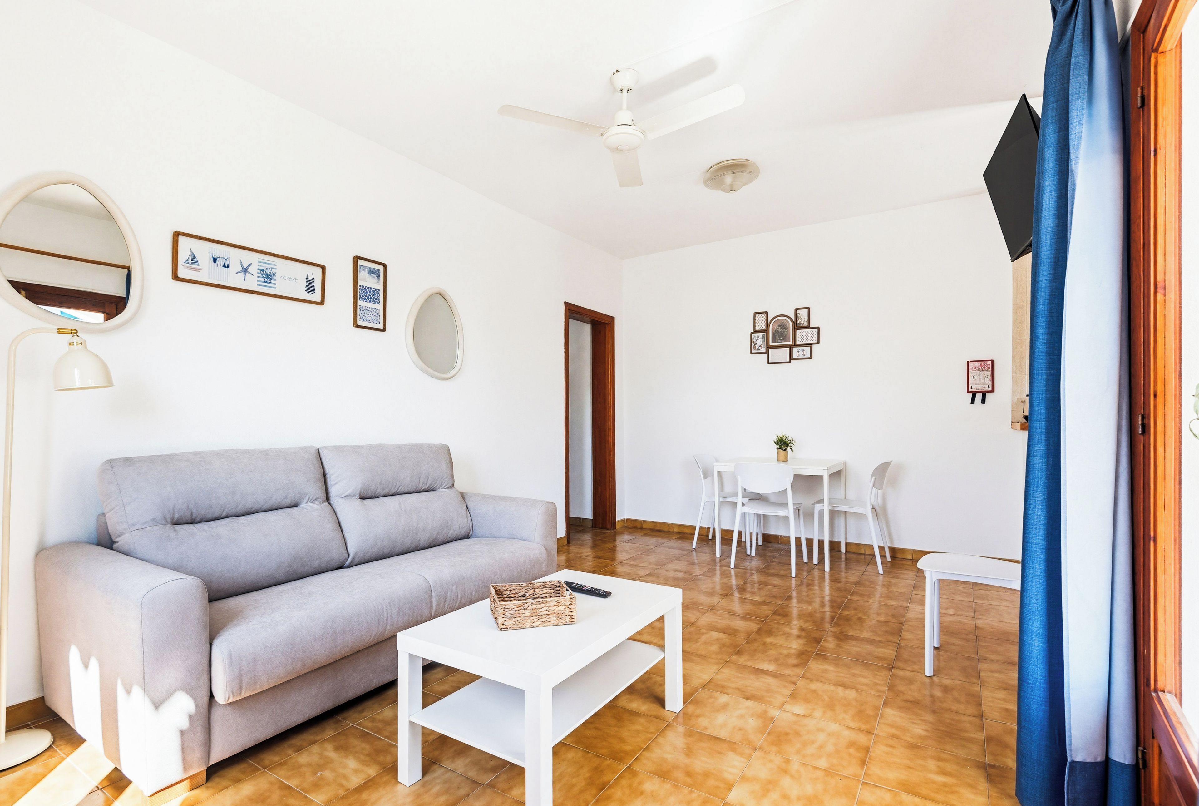 Sala de estar com sofá cinza, mesa de centro branca, ventilador de teto e área de jantar com mesa e cadeiras brancas.