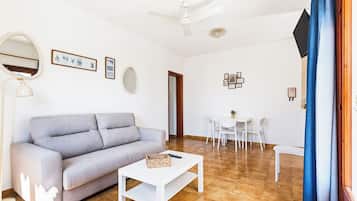 Sala de estar com sofá cinza, mesa de centro branca, ventilador de teto e área de jantar com mesa e cadeiras brancas.