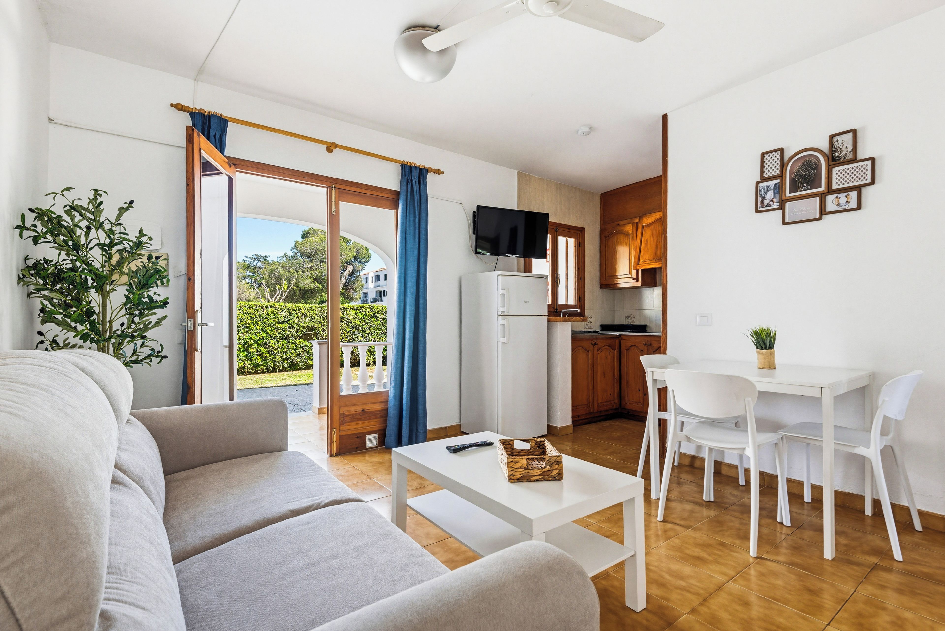 Una sala de estar moderna con un sofá, una mesa de centro y un televisor. Se observa un comedor con una mesa y sillas. Se ve una cocina con armarios de madera y un refrigerador.