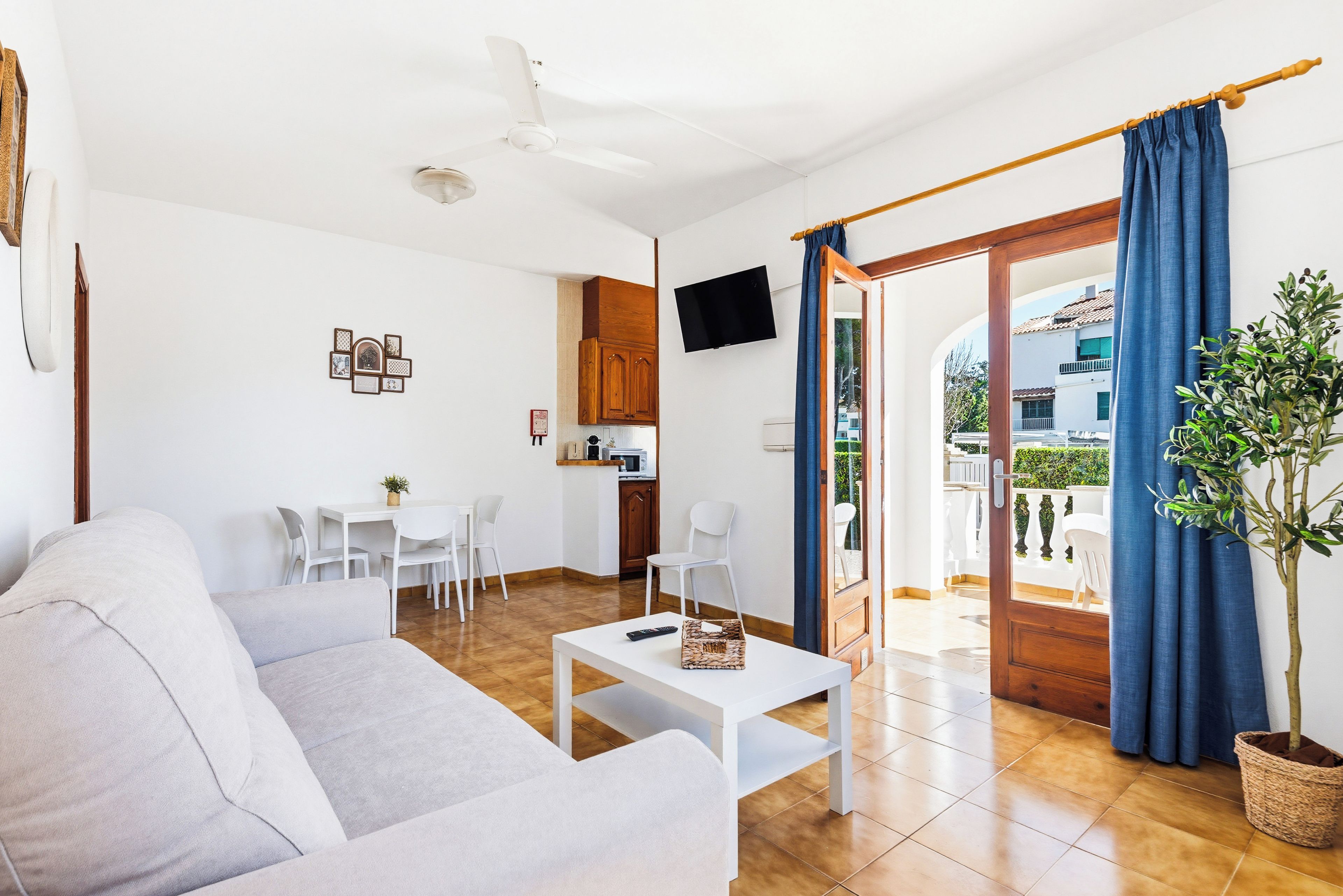 Una sala moderna con un sofá, una mesa de centro y un televisor montado en la pared. Se observa un comedor con sillas y una mesa, además de una cocina con armarios y un microondas.