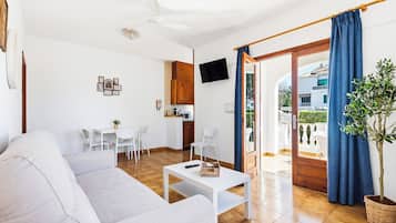 Una sala moderna con un sofá, una mesa de centro y un televisor montado en la pared. Se observa un comedor con sillas y una mesa, además de una cocina con armarios y un microondas.