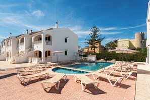 A pool area with lounge chairs, a clear blue sky, and a white building with balconies. - Apartamentos Mar Blanca (Ciutadella de Menorca)