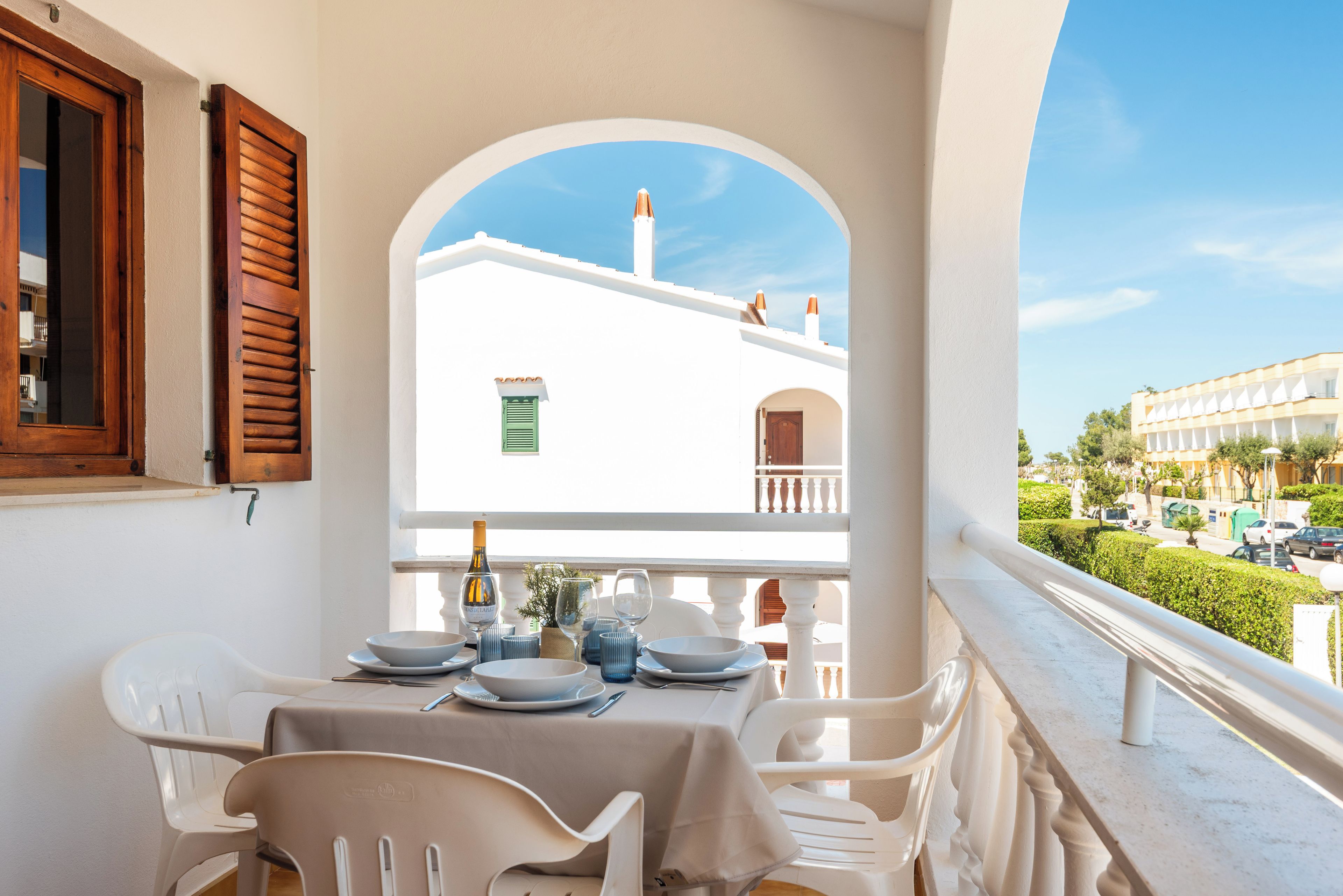 Varanda com mesa posta para refeições, com vista para um edifício de fachada branca e persianas verdes.