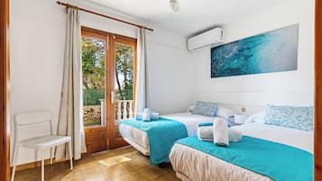 A hotel room with two beds, a chair, an air conditioning unit, and a view of trees through a door.