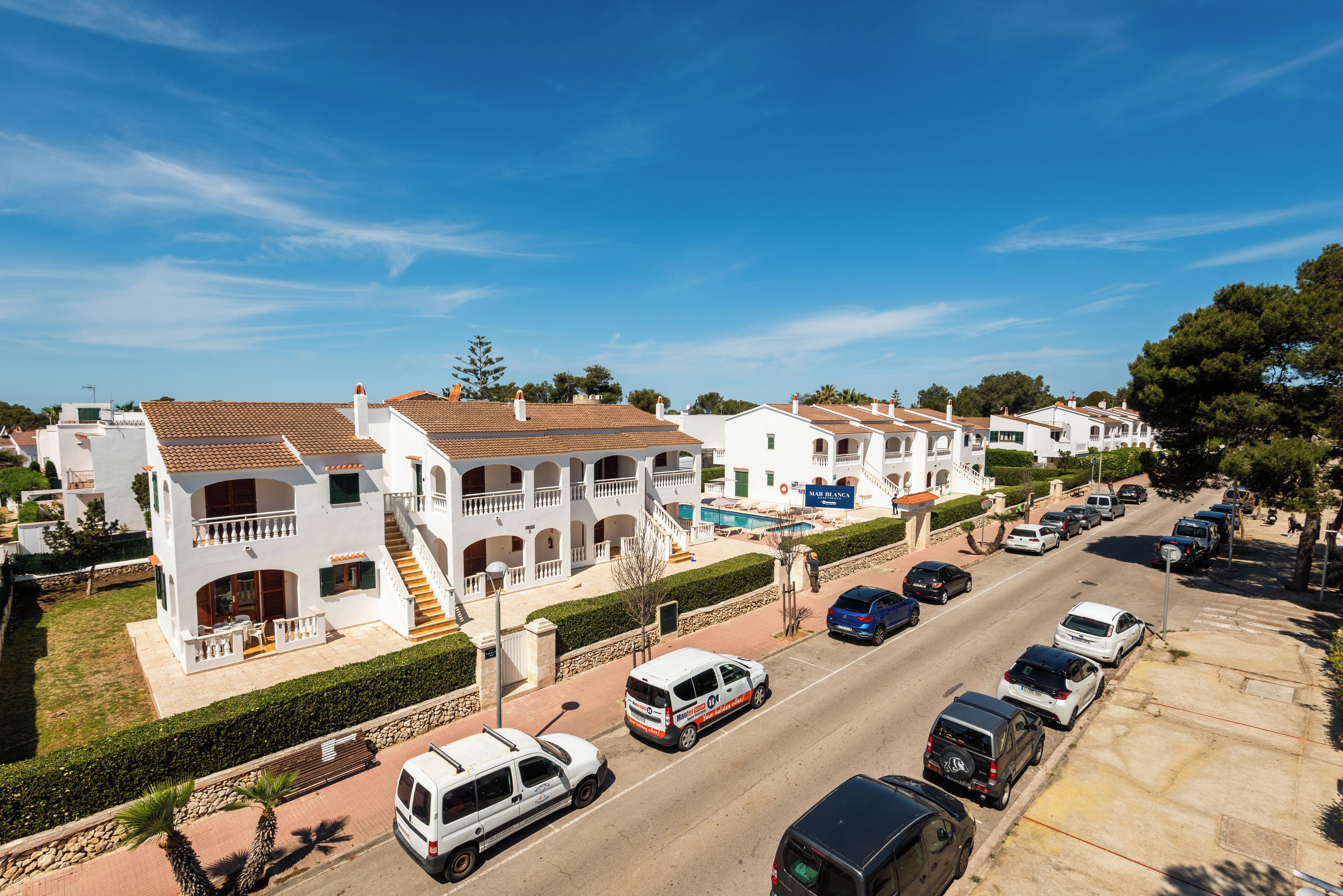 A residential street with white houses, a swimming pool, and parked cars.