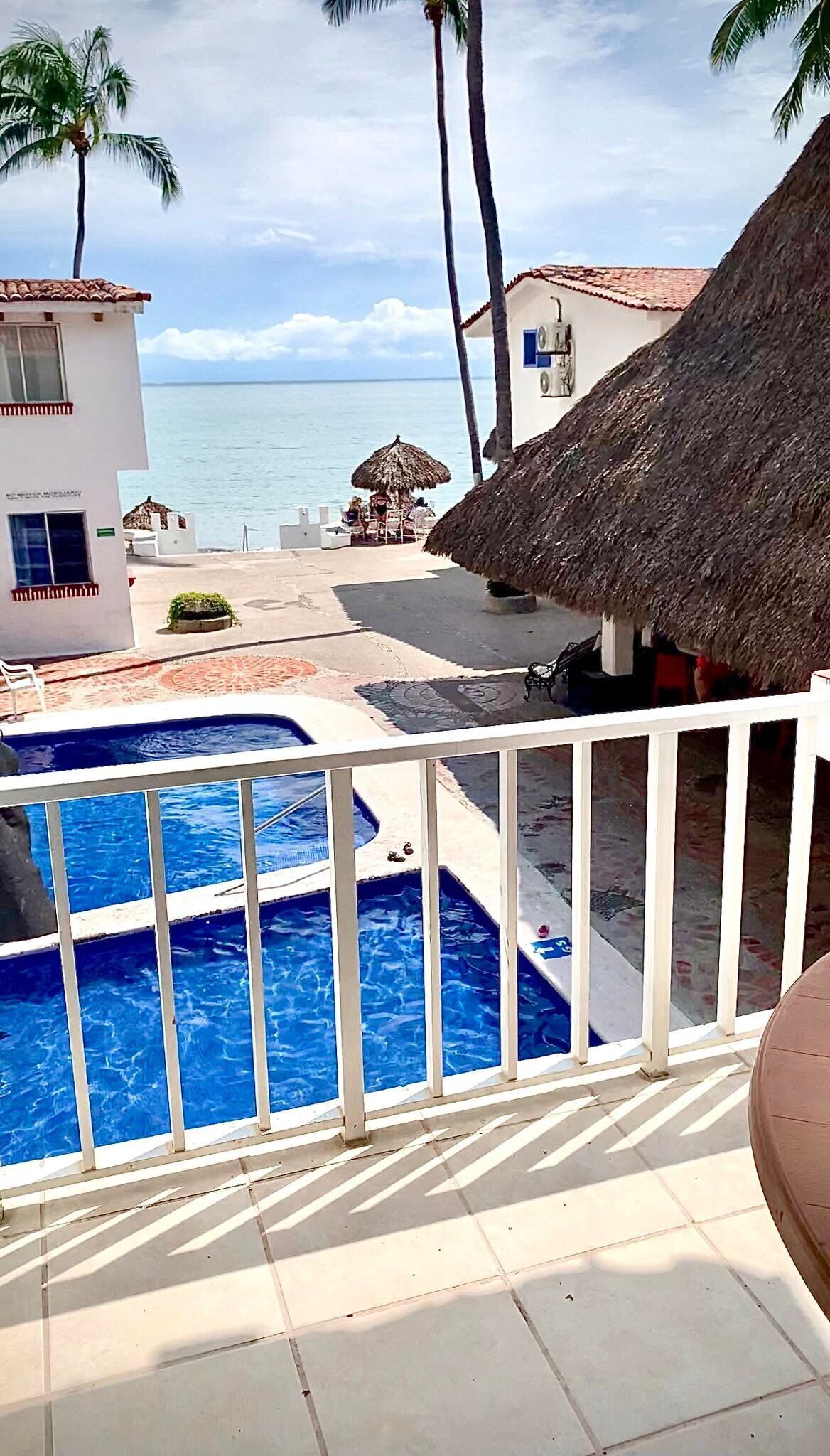 A pool area with a view of the sea, a thatched-roof structure, and a building with a balcony.