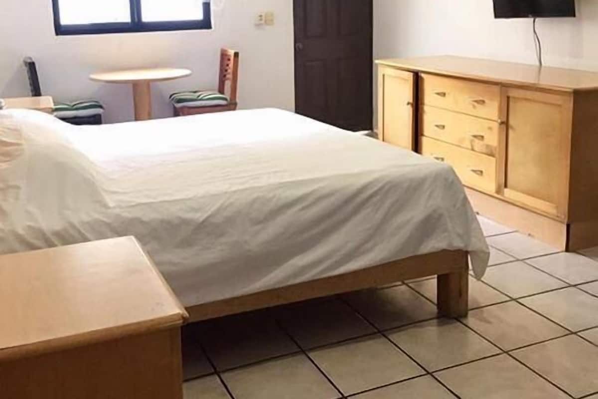 A bedroom with a bed, a wooden dresser, a television mounted on the wall, and a window with blinds.