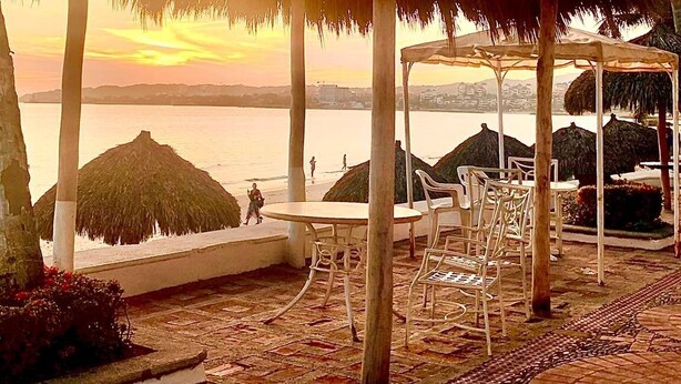 Un patio frente a la playa con un toldo de paja, sillas de descanso y vista al océano.