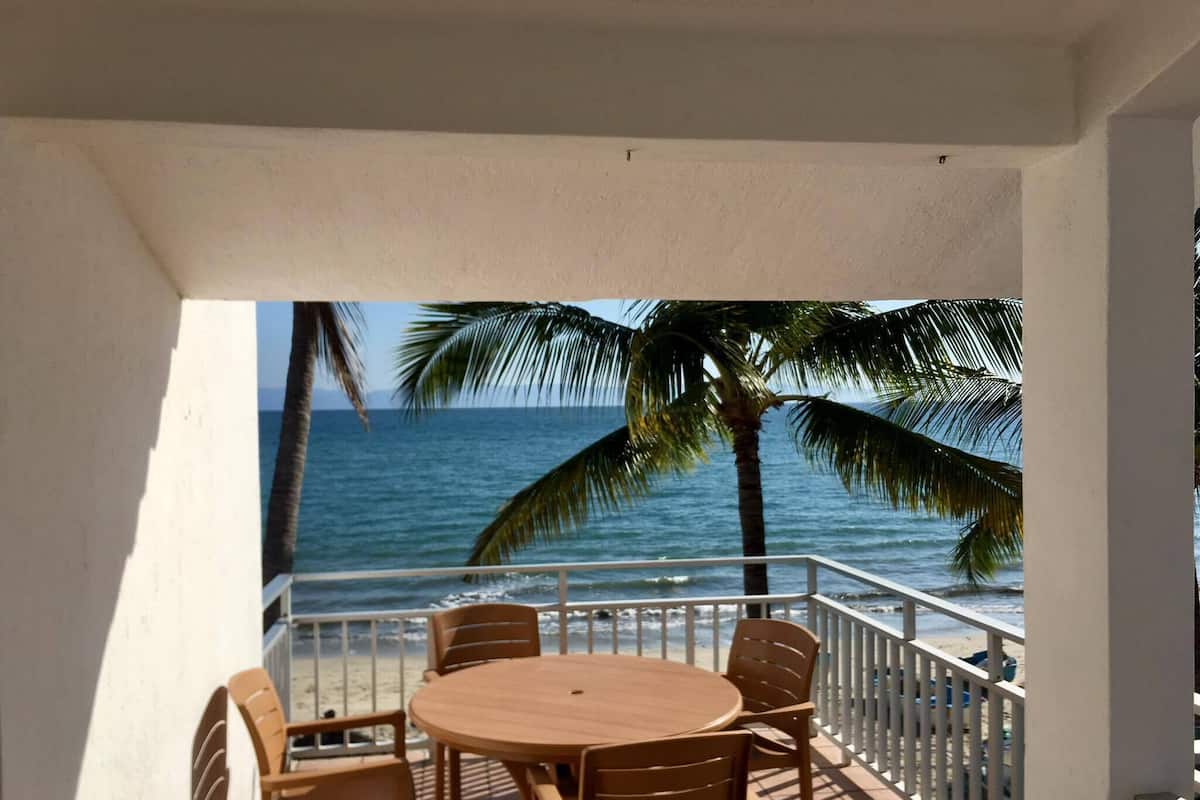 A balcony with a round wooden table and chairs, overlooking a beach with palm trees and the ocean.