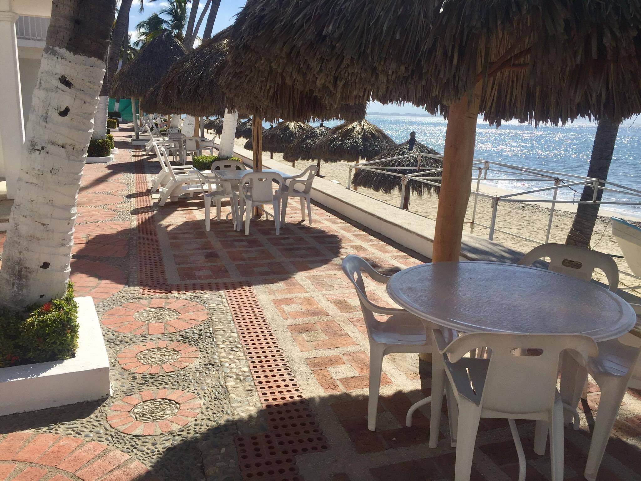 Un patio frente a la playa con sillas y mesas de plástico blanco, sombrillas de paja y vista al océano.