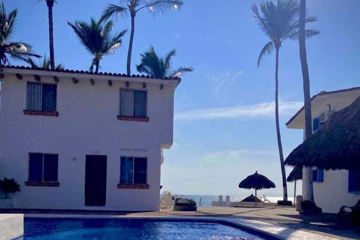 A swimming pool with a tiled deck, a white building with multiple windows, and palm trees in the background.