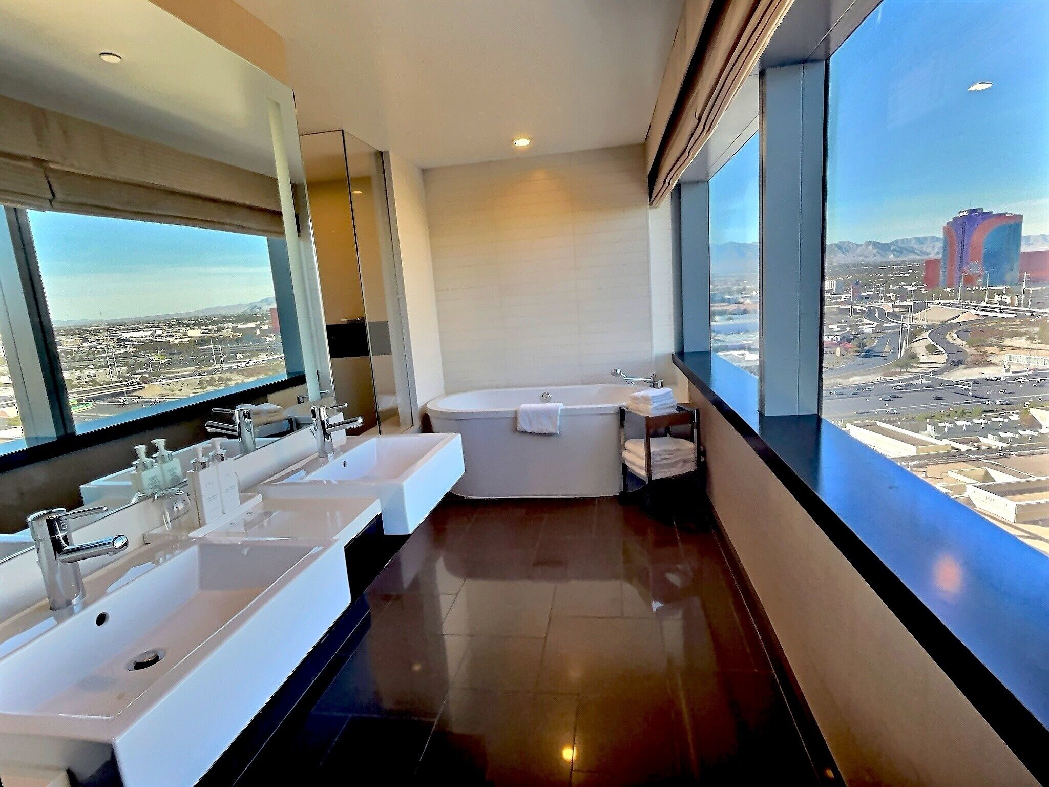A modern bathroom with dual sinks, a bathtub, and a large window offering a city view.