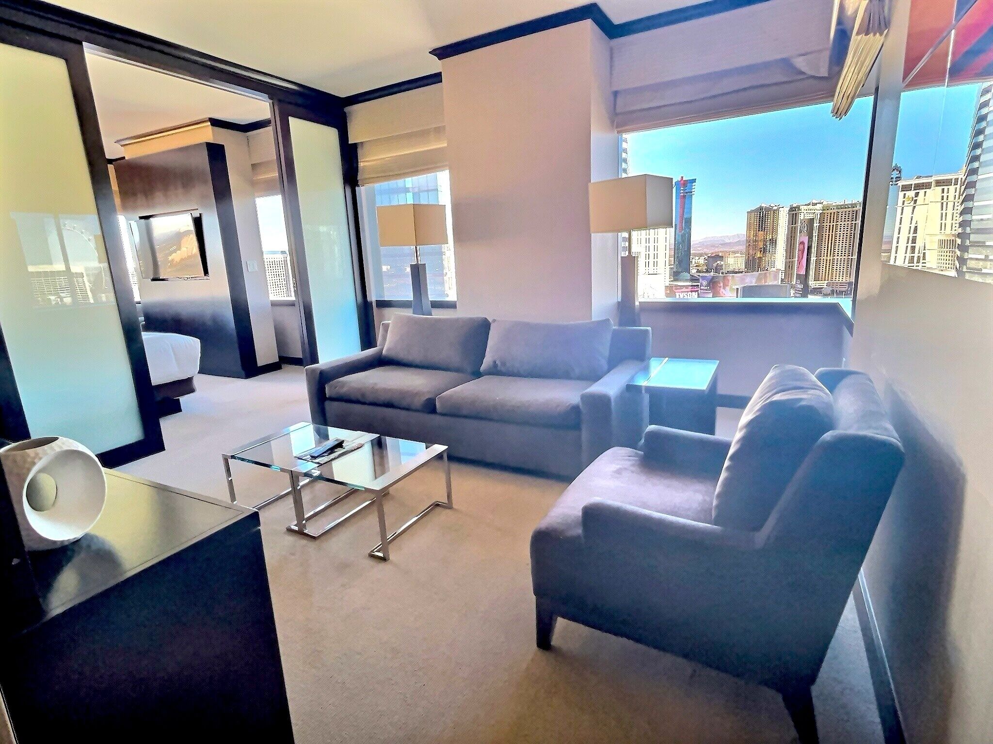 A modern hotel room with a grey sofa, a glass coffee table, and a view of the cityscape through large windows.