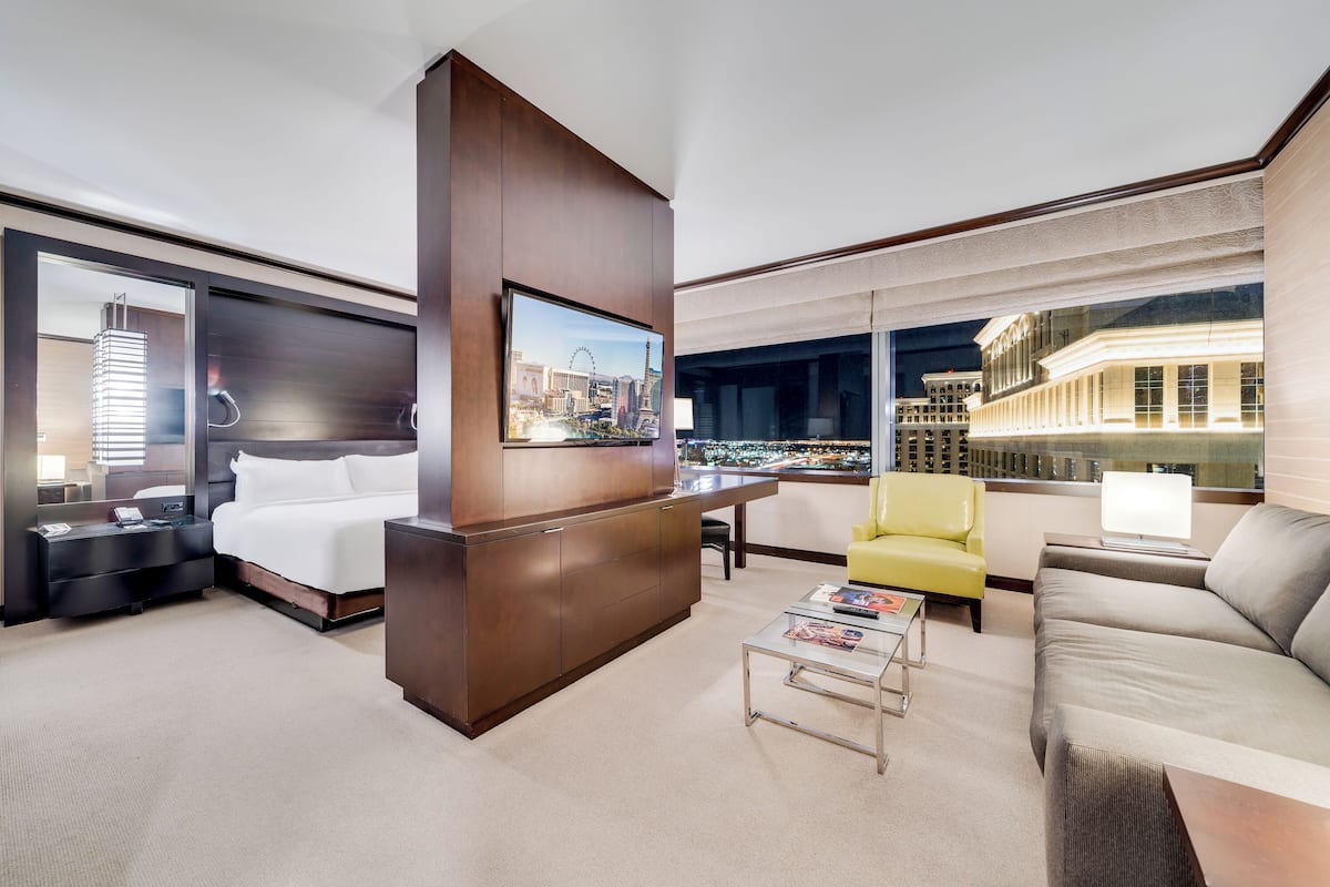 A modern hotel room with a large bed, a desk, a yellow armchair, a grey sofa, and a city view through large windows.