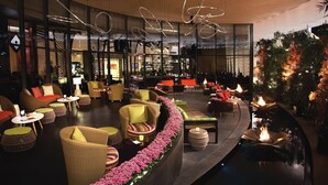 A spacious indoor lounge with wicker furniture, a central fire feature, and a view of the bar area through large windows.