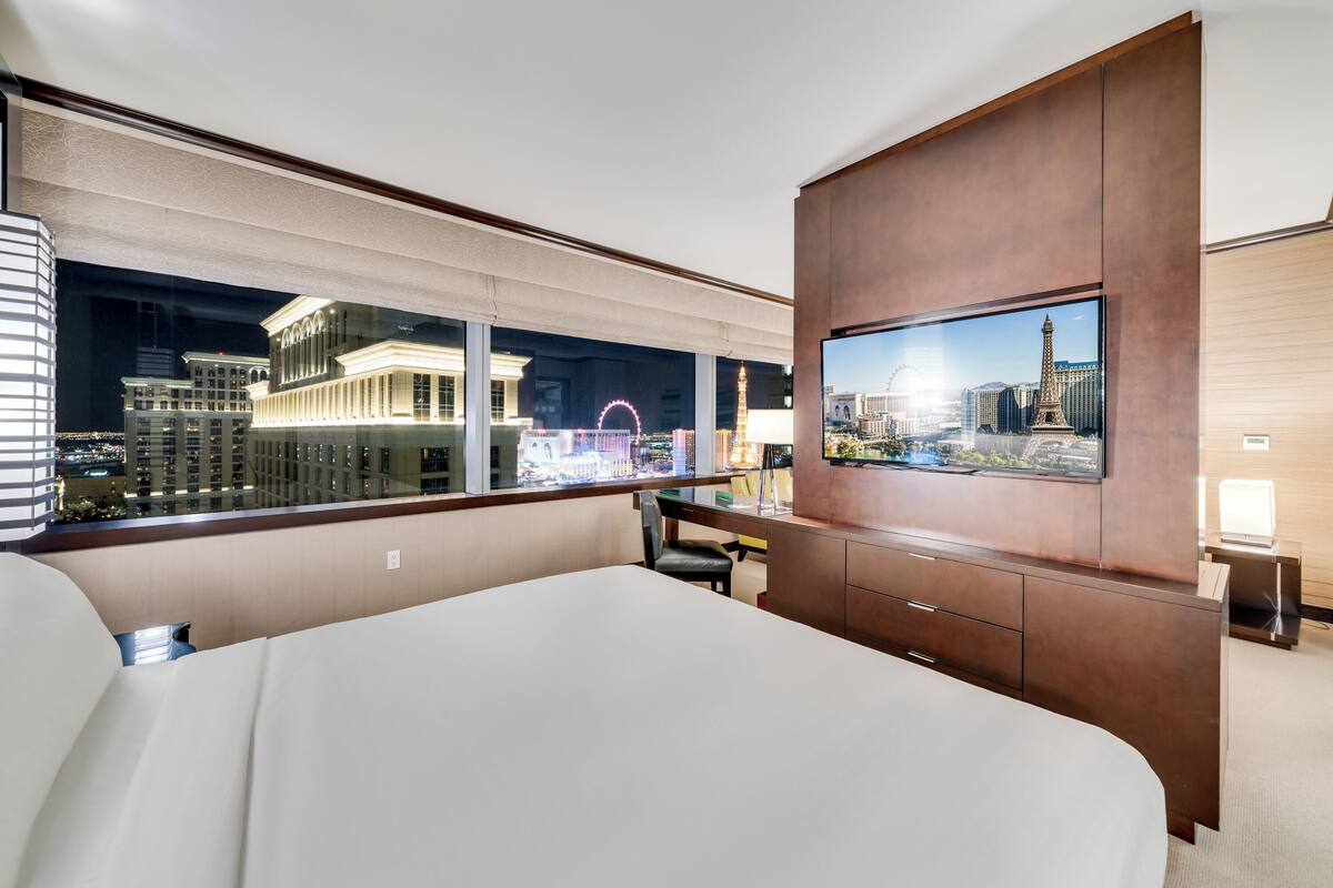 A hotel room with a large bed, a TV mounted on a wooden cabinet, a desk with a chair, and a view of a cityscape including the Eiffel Tower.