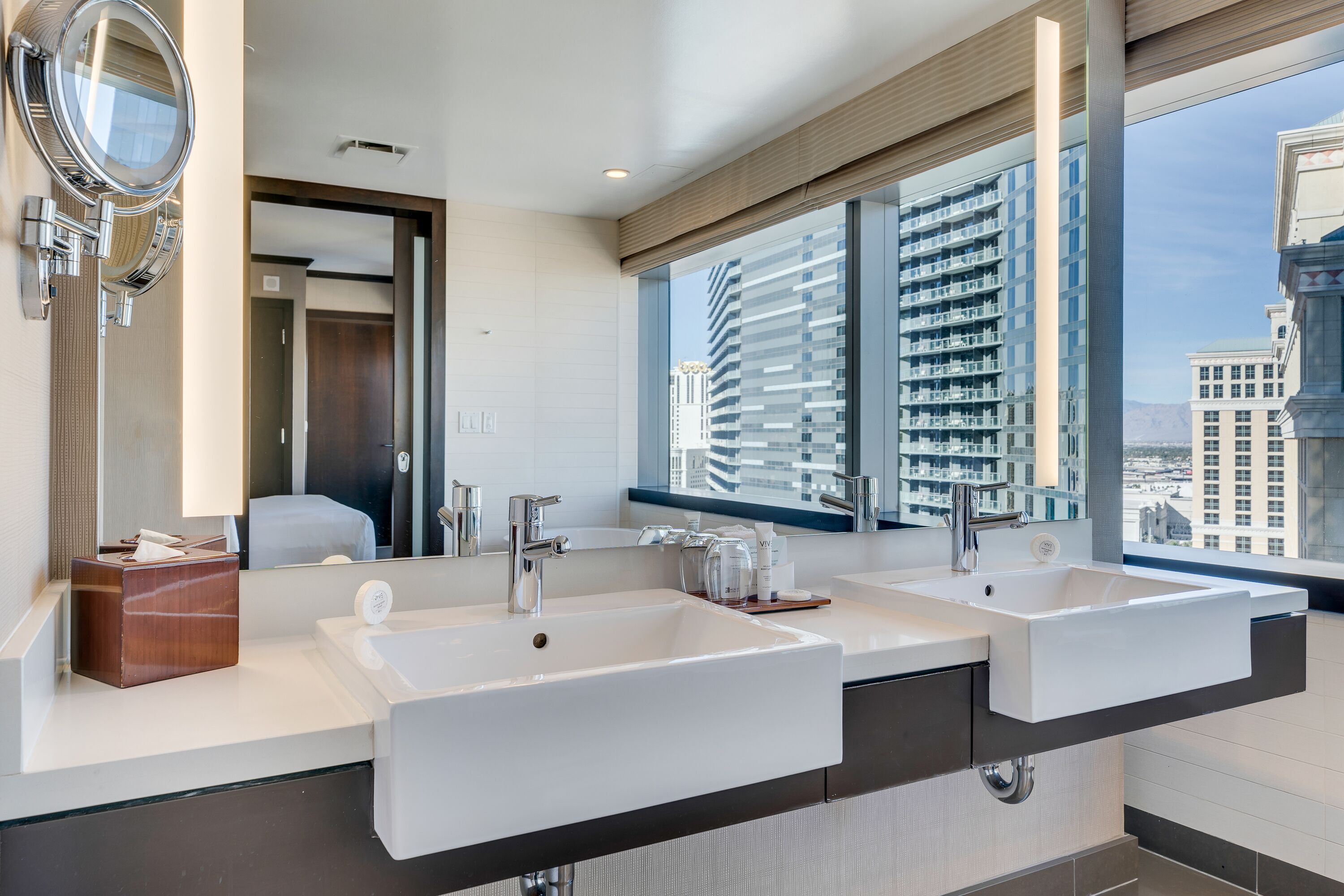 A modern bathroom with two sinks, a large mirror, and a view of tall buildings.