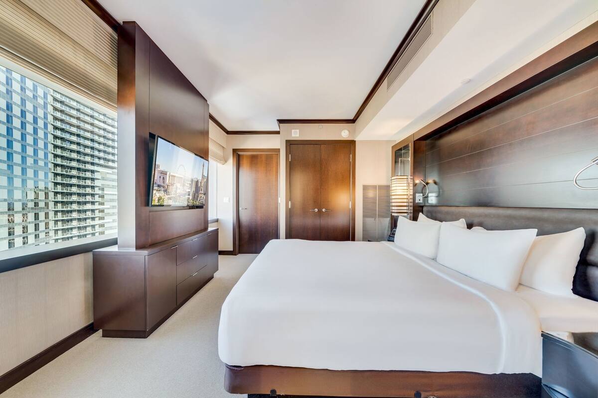 A hotel room with a large bed, a TV, a view of a cityscape, and a wooden headboard.