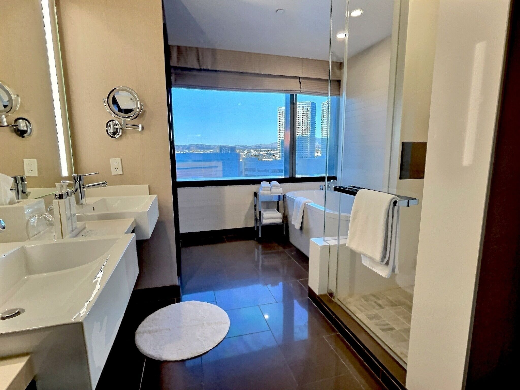 A modern bathroom with dual sinks, a bathtub, and a large window offering a city view.