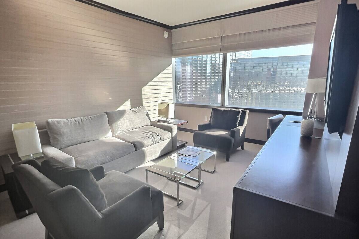 A modern living room with a grey sofa, armchairs, a coffee table, and a TV on a stand. Large windows offer a view of the cityscape.
