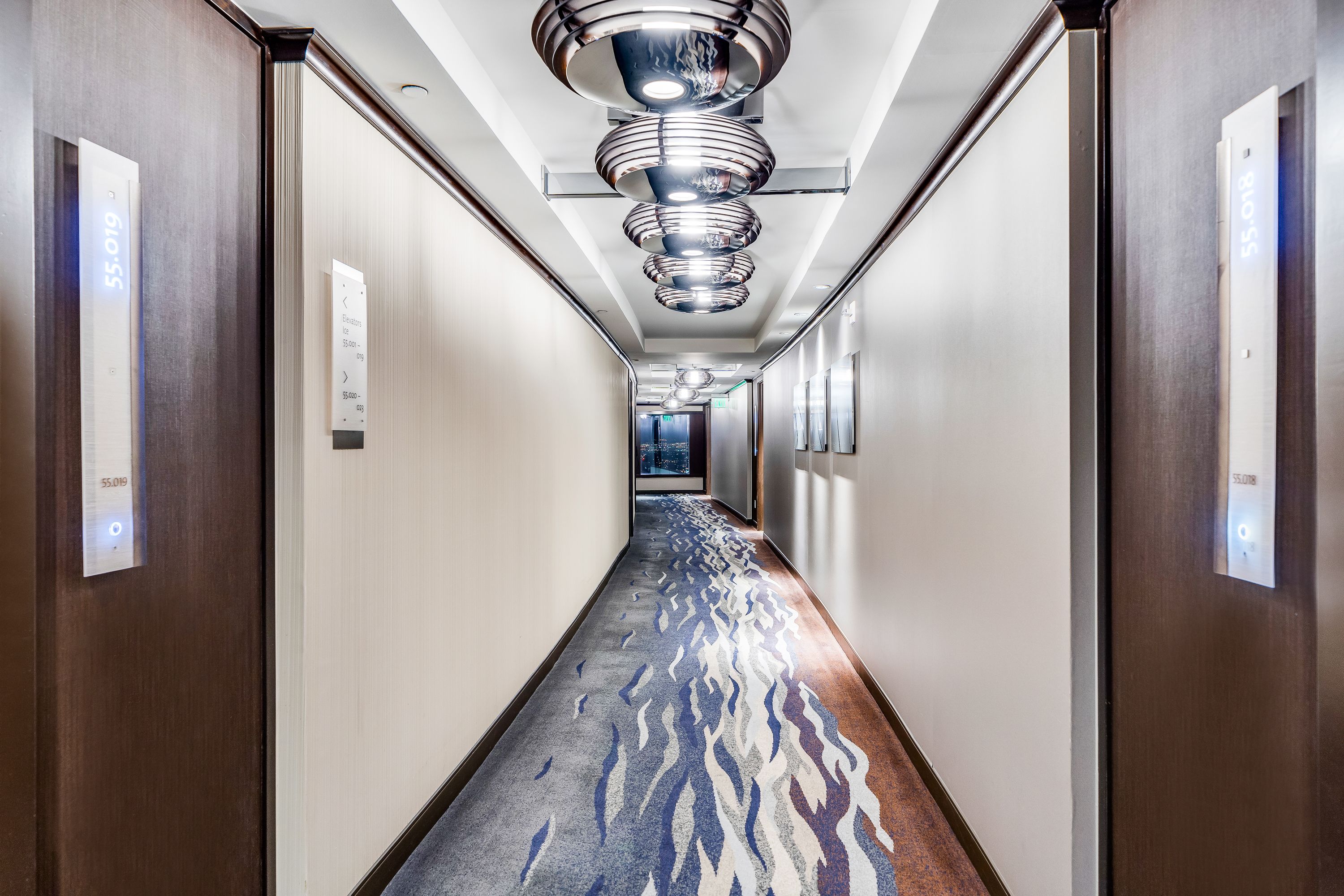 A modern hotel hallway with a patterned carpet, wooden walls, and a ceiling with circular light fixtures.