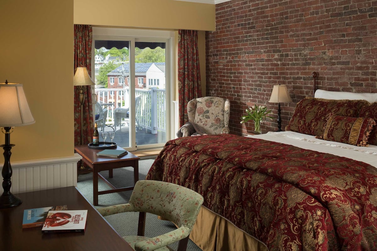 Una habitación de hotel con una cama grande, una mesita de noche de madera, una silla, una mesa con lámpara y vistas al exterior.