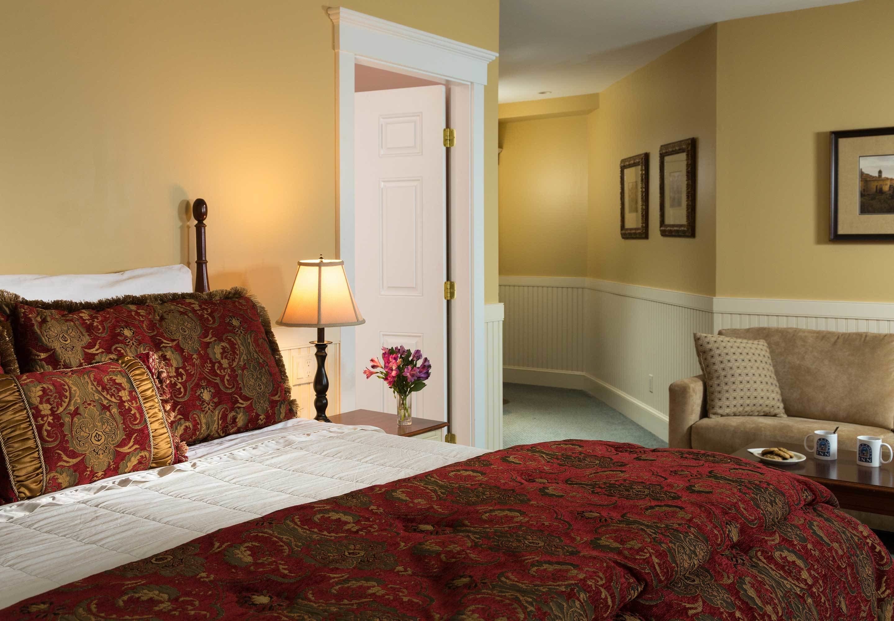 A hotel room with a large bed, a bedside table with a lamp, a vase with flowers, and a sofa with a plate and mug.