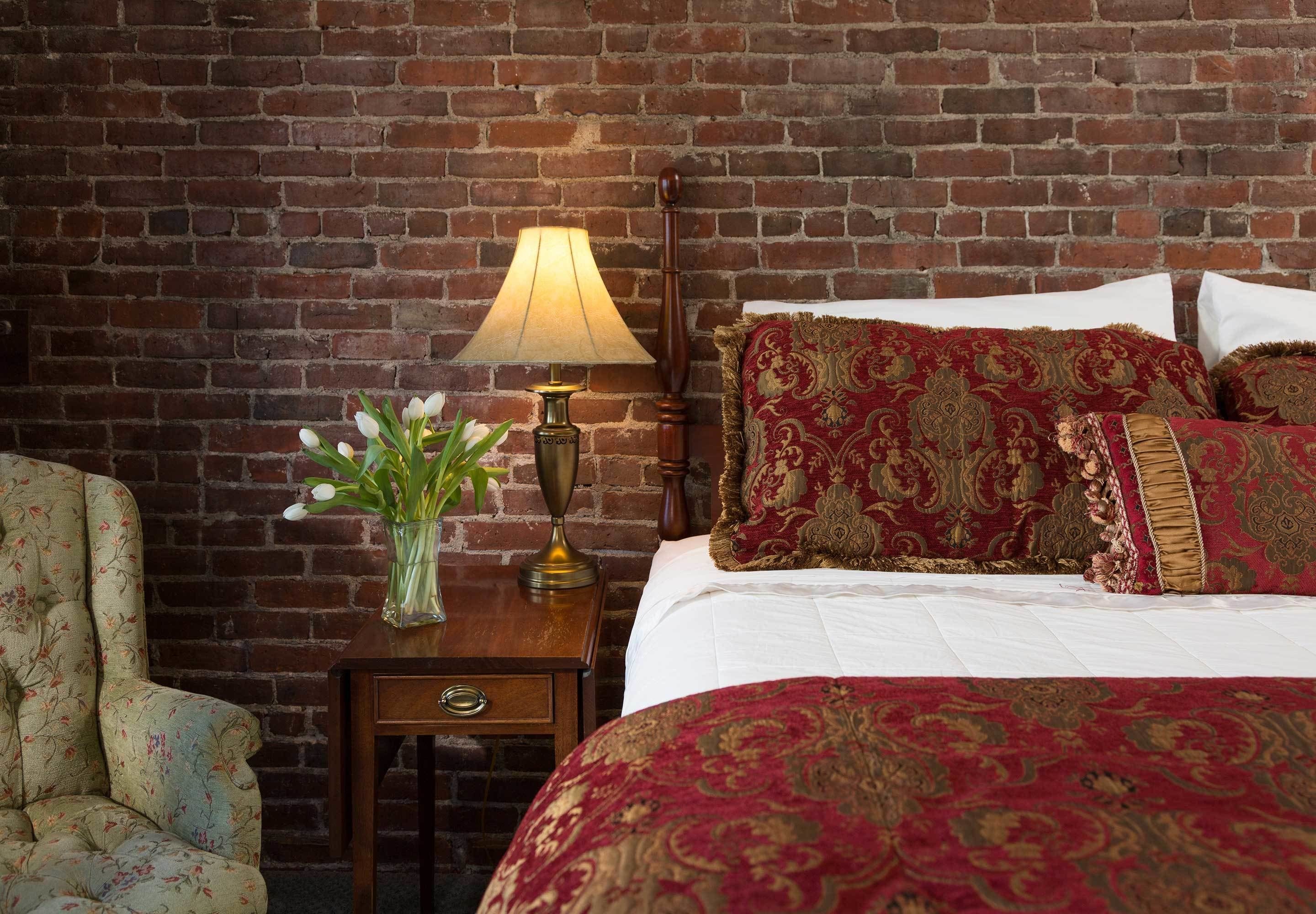 Una habitación de hotel con pared de ladrillo, mesita de noche de madera con lámpara, sillón floral y cama con ropa de cama roja y dorada.