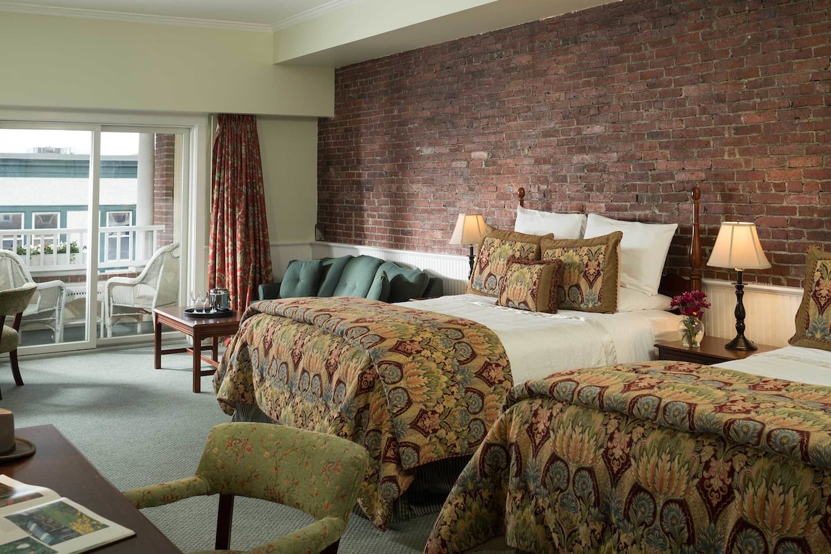 Habitación de hotel con pared de ladrillo, dos camas con ropa de cama estampada, un balcón con sillas y una mesa con lámpara.