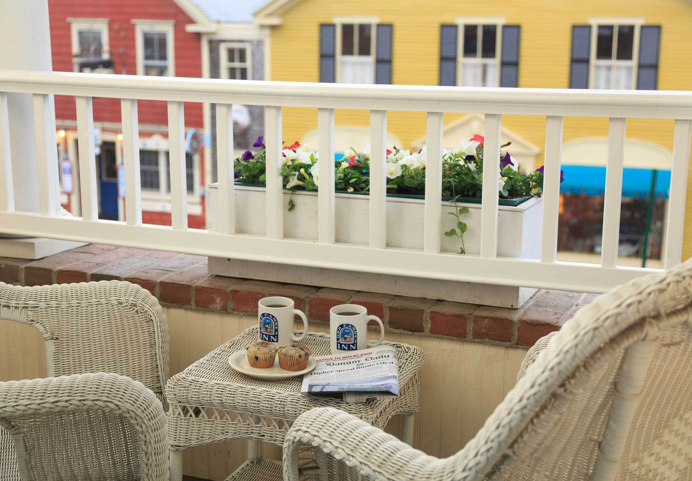 A balcony with wicker chairs, a small table with two coffee mugs and a plate of muffins, and flower boxes with colorful flowers.