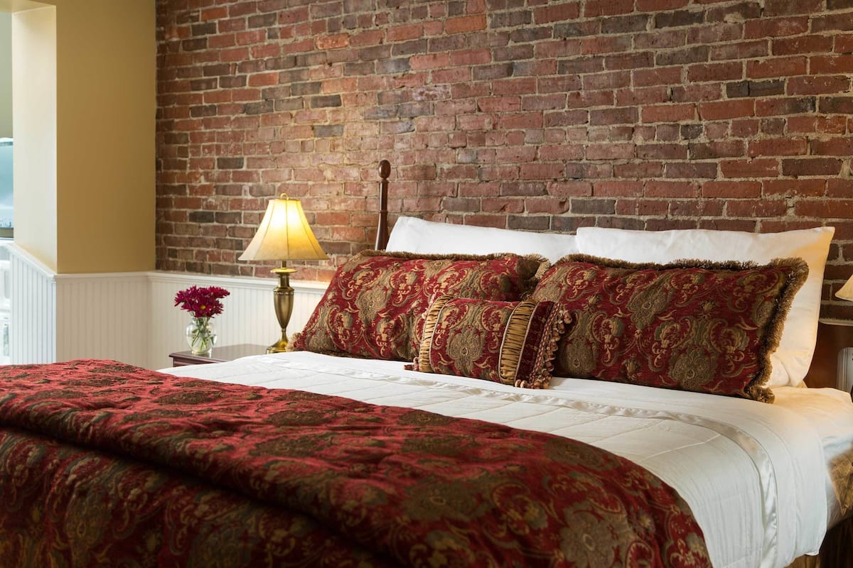 Una cama con una colcha de motivos dorados y rojos, almohadas blancas y un cabecero de madera frente a una pared de ladrillo.