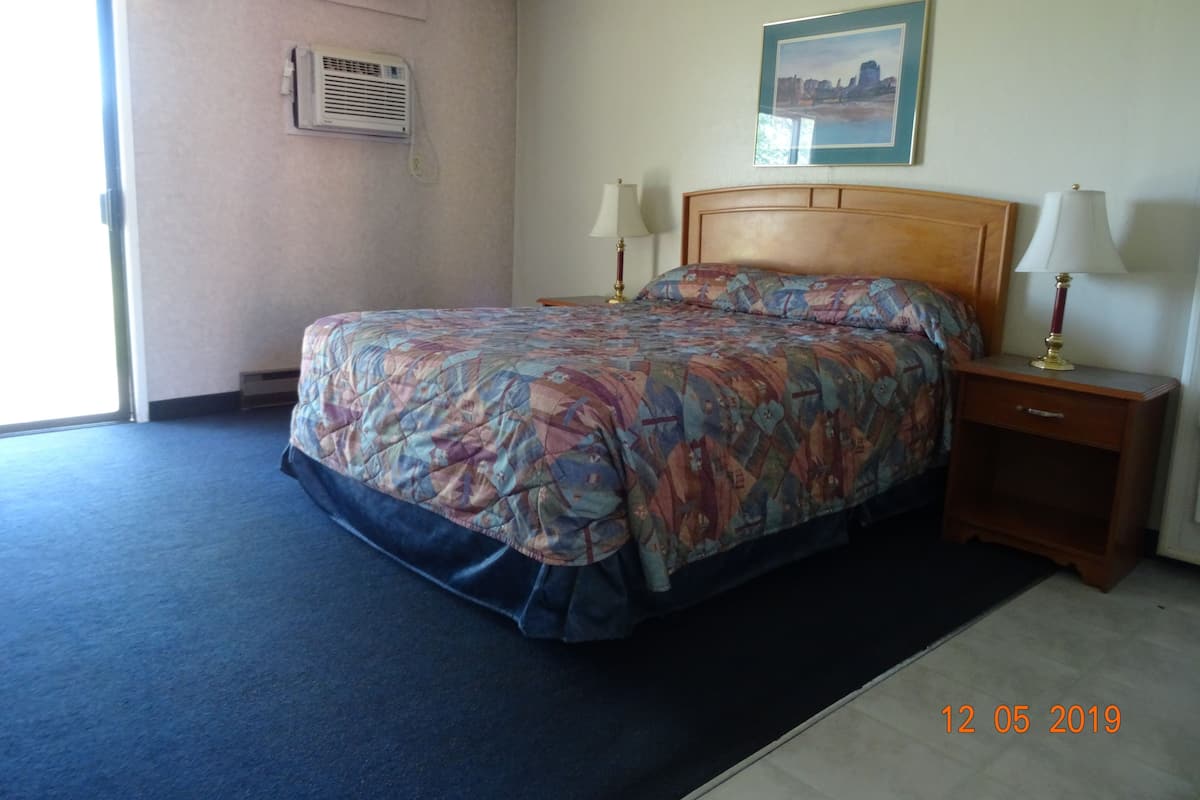 A hotel room with a bed, bedside lamps, a nightstand, a wall-mounted air conditioner, and a framed picture on the wall.