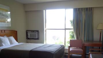 A hotel room with a bed, a desk, a chair, a window with curtains, and an air conditioning unit.
