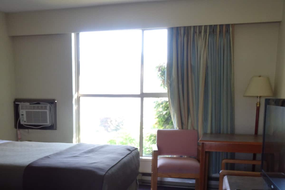 A hotel room with a bed, a desk, a chair, a window with curtains, an air conditioning unit, and a painting on the wall.