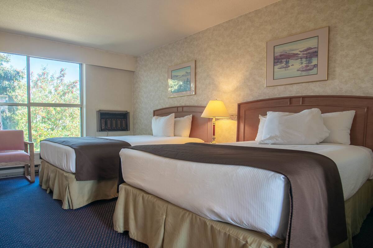 A hotel room with two beds, a window with a view of trees, a lamp, and a painting on the wall.