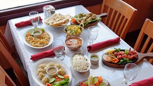 Een eettafel gedekt met diverse gerechten, waaronder pasta, rijst en gegrild vlees, met houten stoelen eromheen.