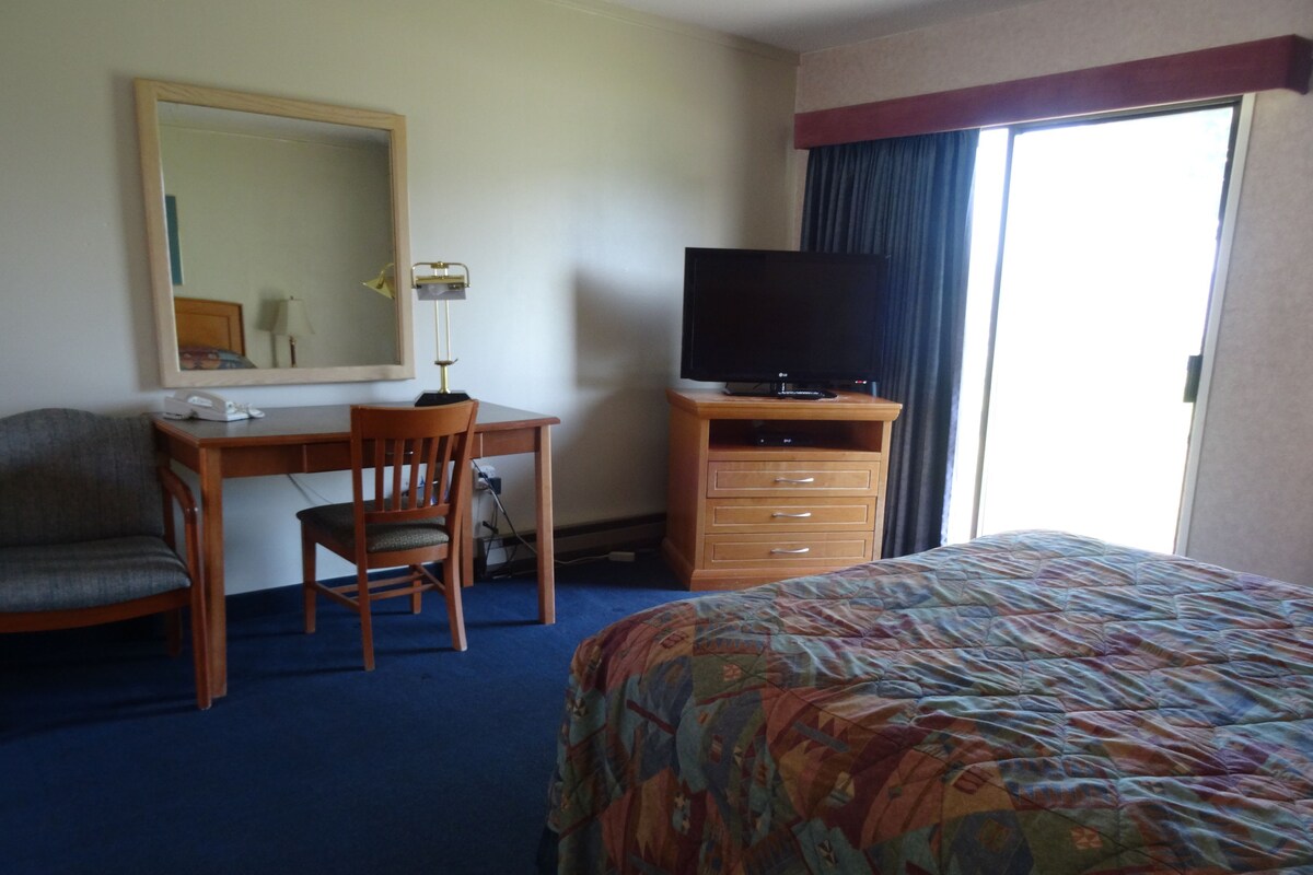 A hotel room with a bed, a desk with a chair, a television on a stand, a mirror, and a window with curtains.