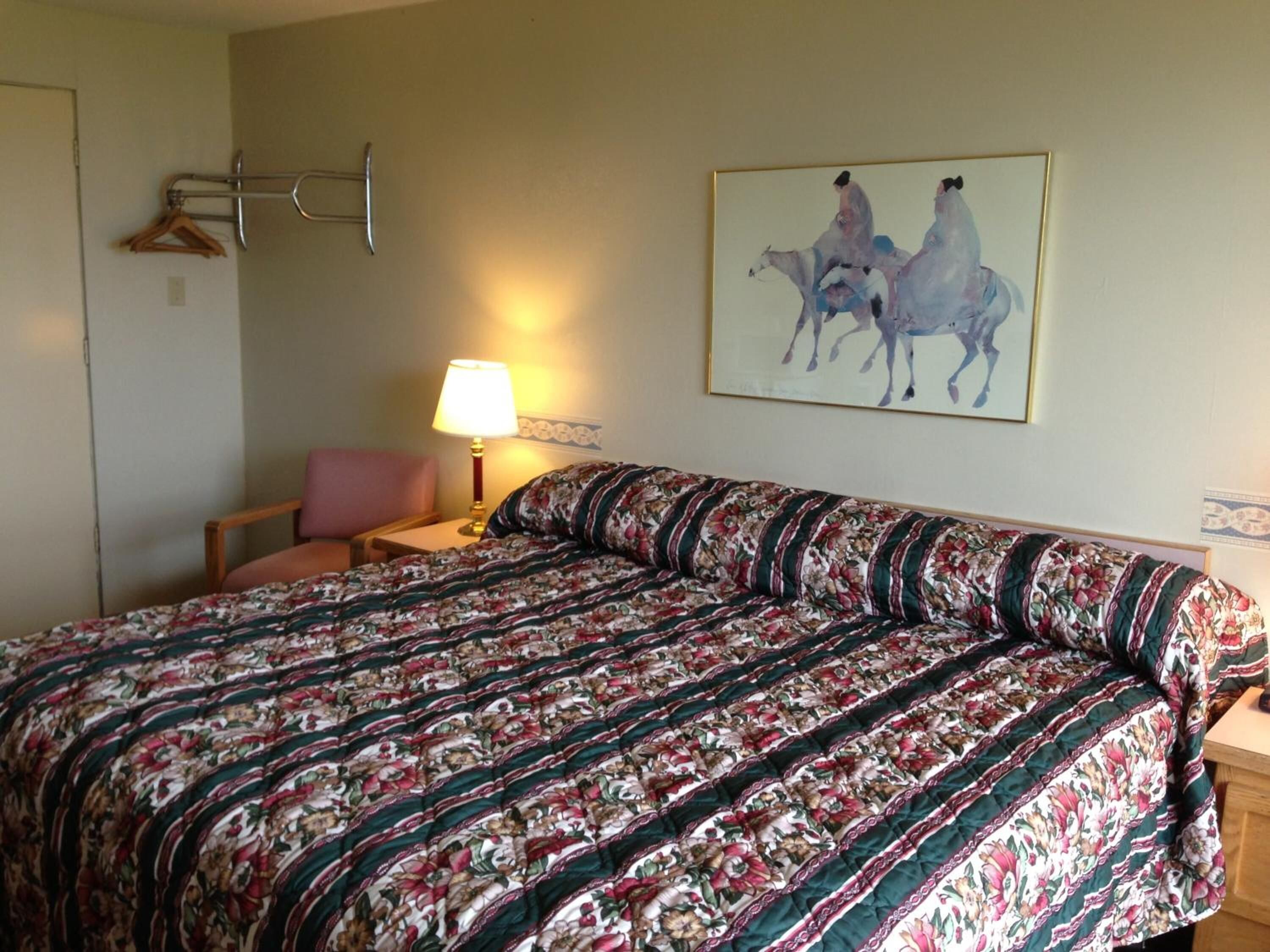 A hotel room with a large bed, a chair, a bedside table with a lamp, and a wall-mounted coat rack.