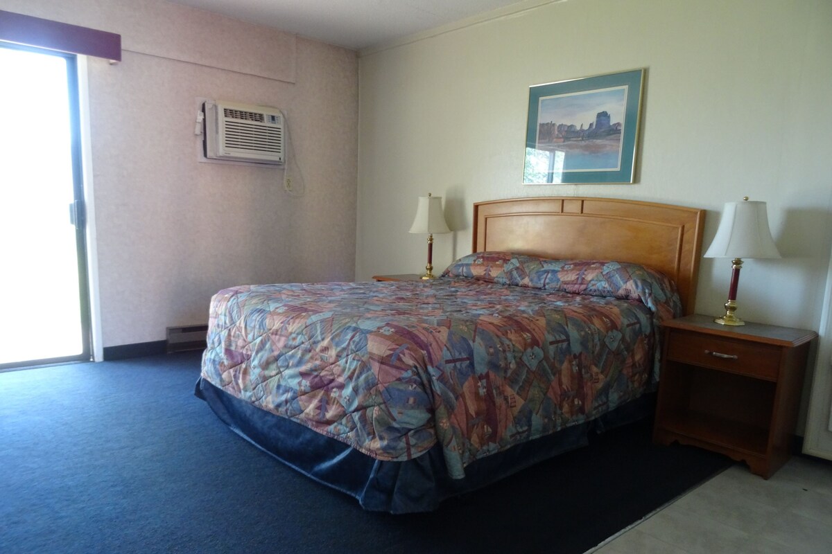 A hotel room with a bed, two bedside lamps, a framed picture on the wall, and an air conditioning unit.