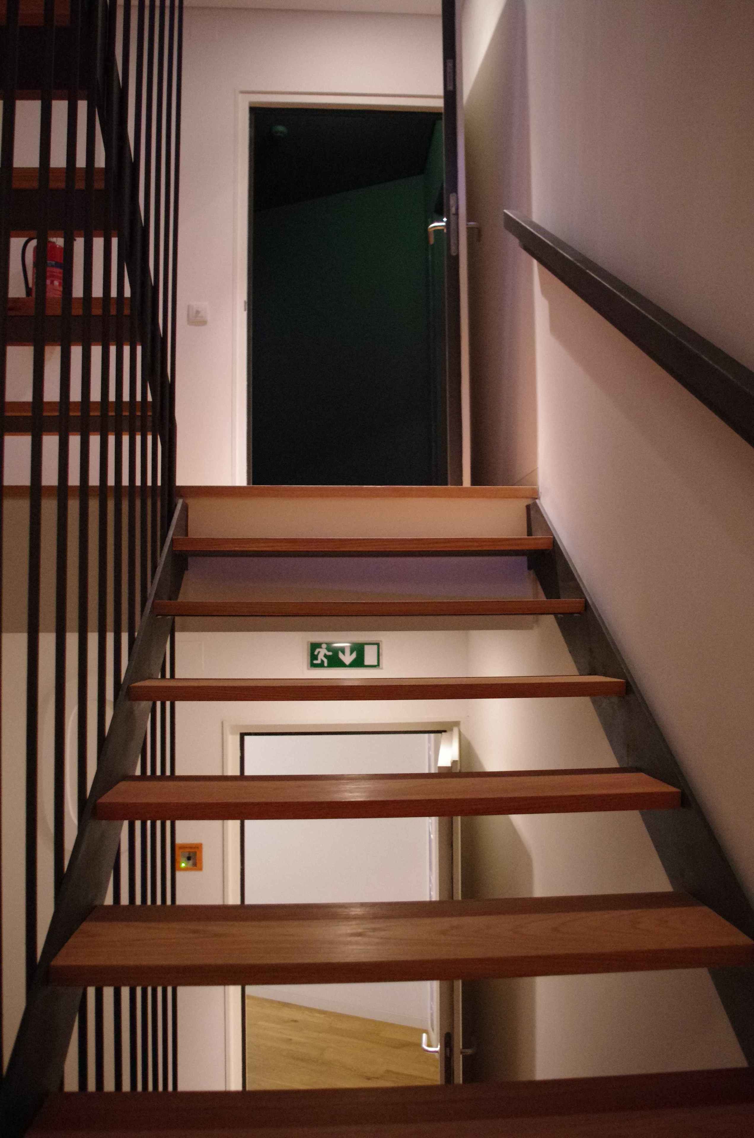 Una escalera con escalones de madera y barandillas de metal, que lleva a una puerta con un letrero de salida de emergencia.