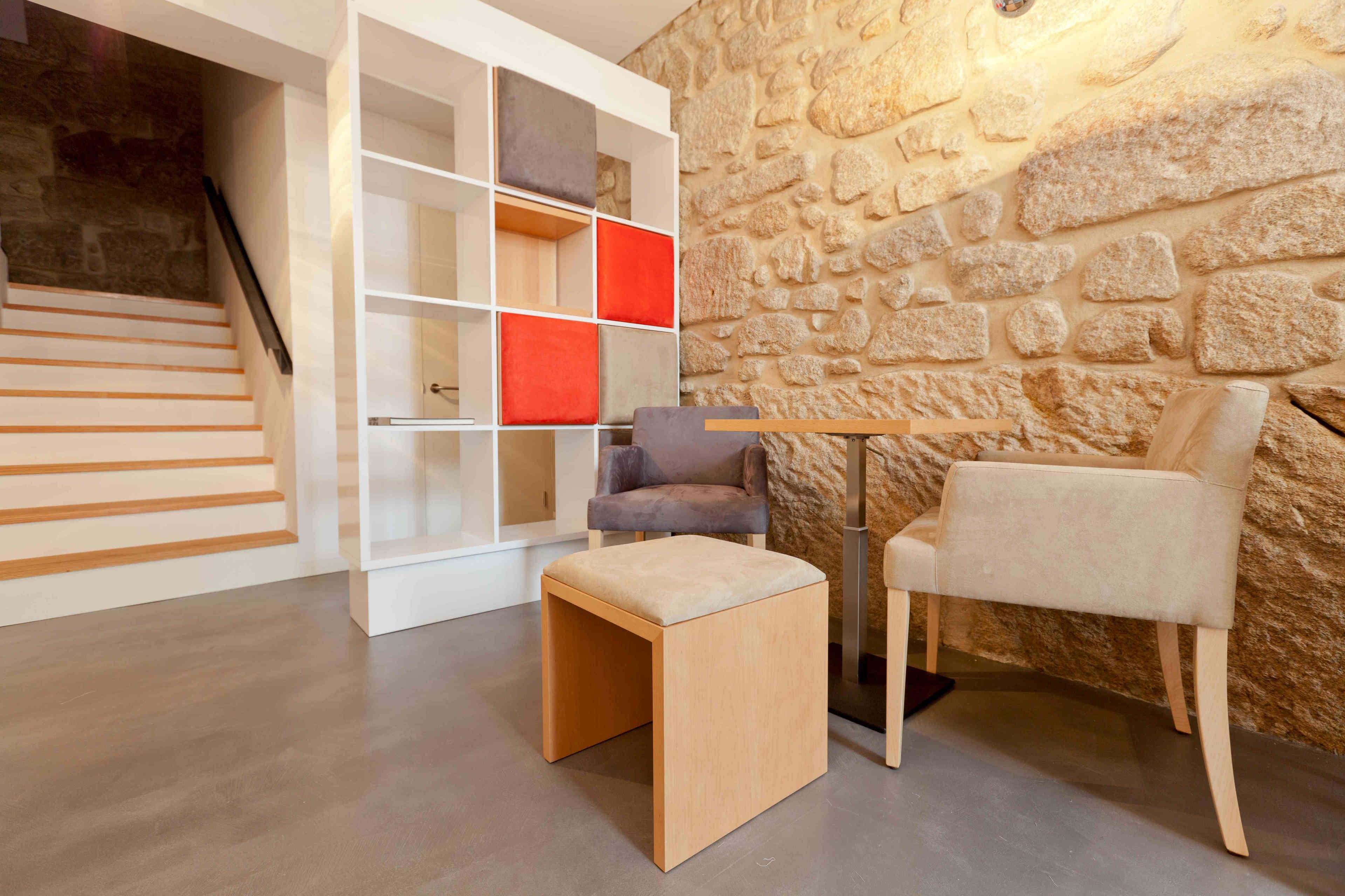A modern interior with a stone wall, a wooden staircase, a white shelving unit with red cushions, a grey chair, and a wooden stool.