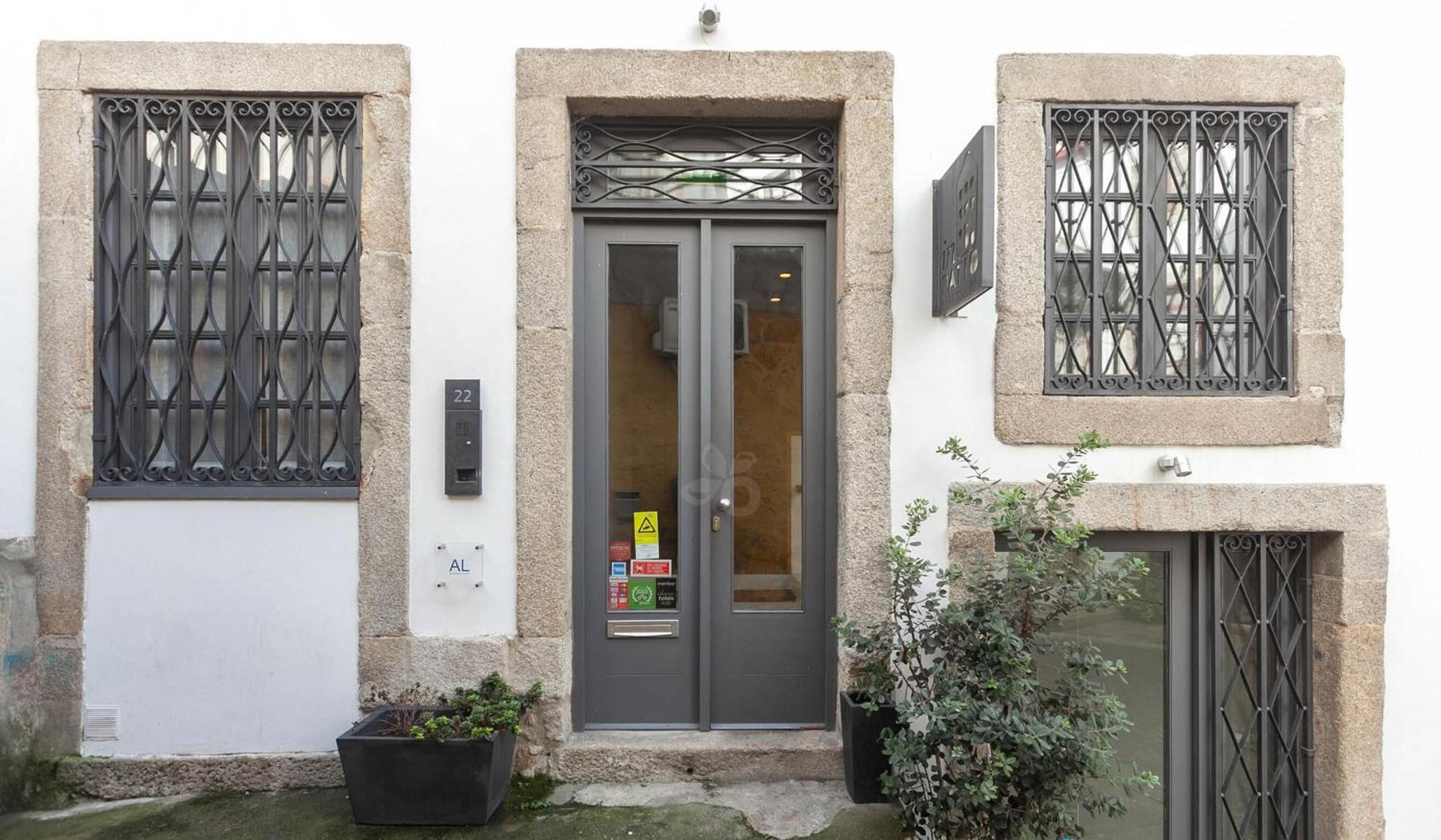 A building with a grey door, two windows with metal bars, and a small planter with plants.