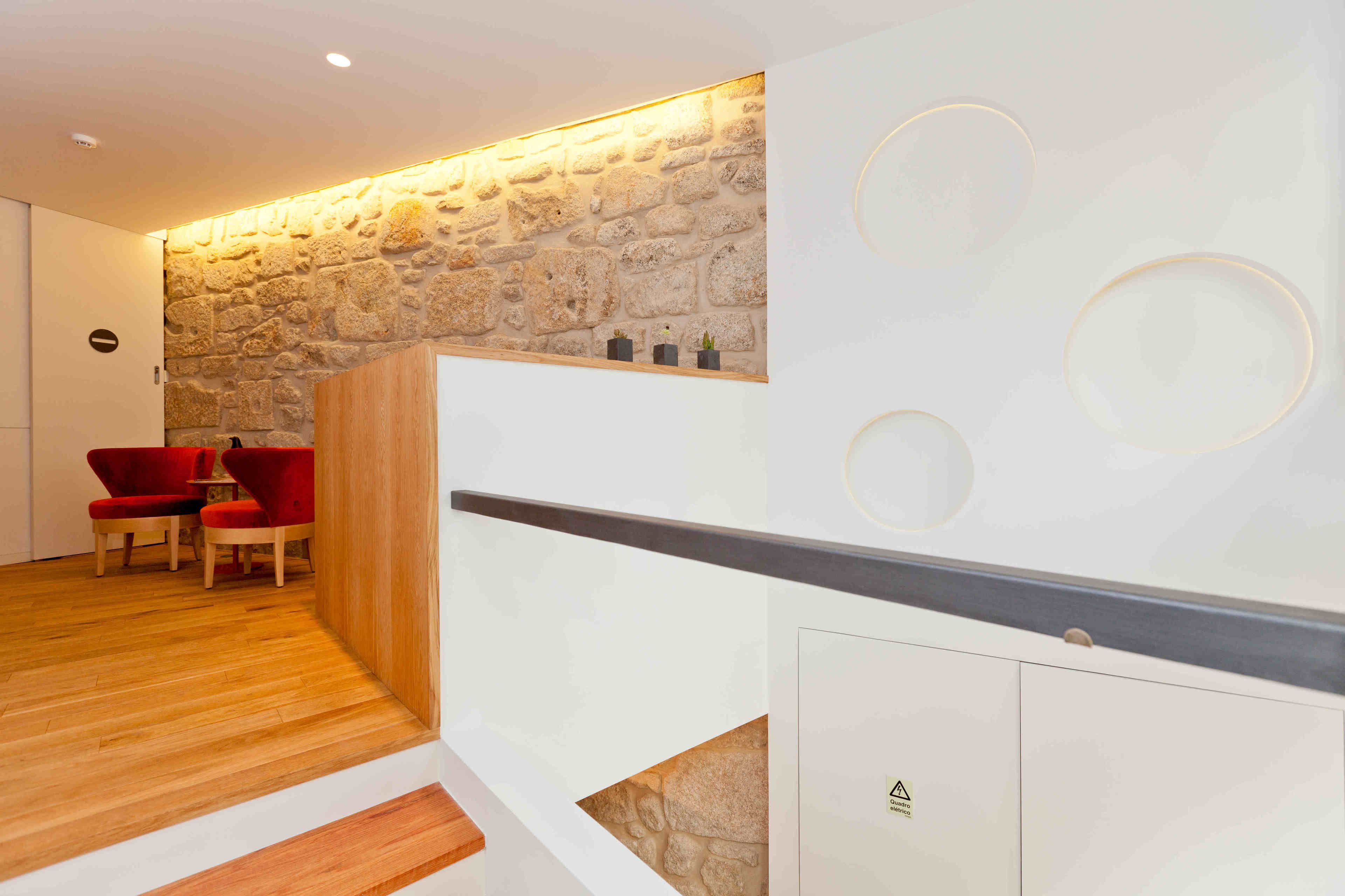 A modern hotel lobby with a stone wall, wooden flooring, and red chairs.