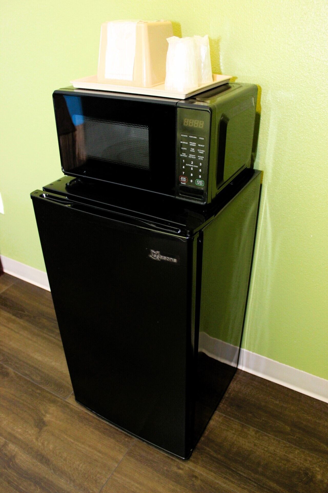 A black refrigerator with a microwave on top, both placed against a green wall.