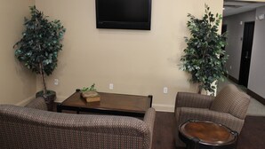 A room with a flat-screen TV mounted on the wall, two potted plants, a wooden coffee table, and two patterned sofas. - Scottish Inns Killeen near Fort Cavazos (Killeen)